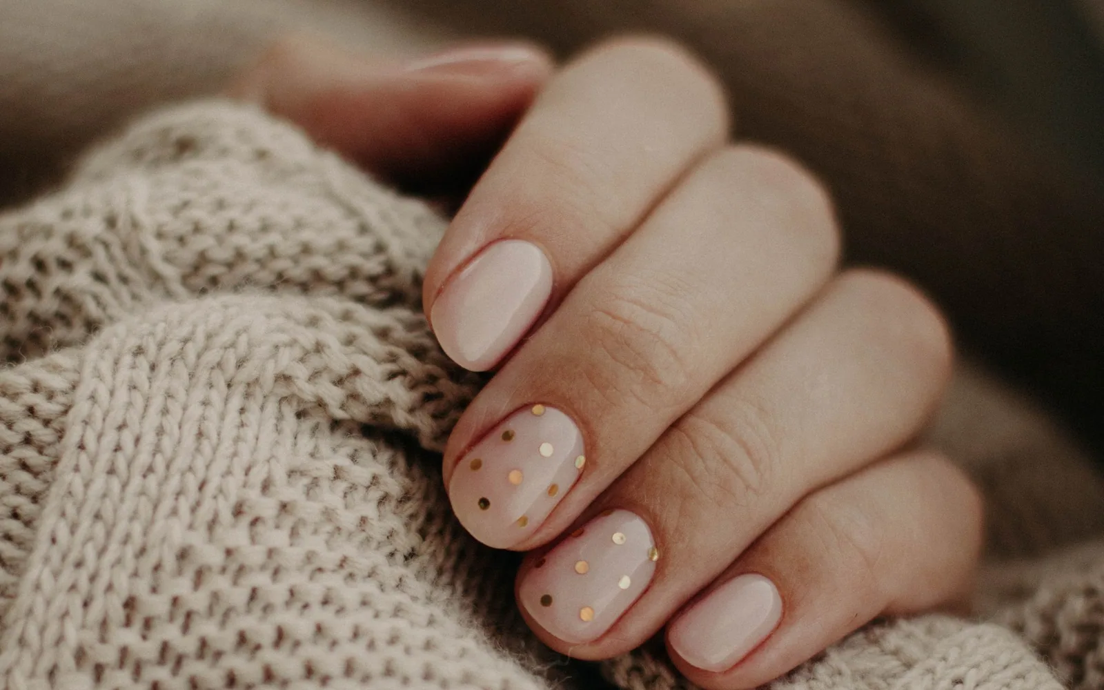 Mãos em repouso com unhas em tom nude sobre tecido de tricô